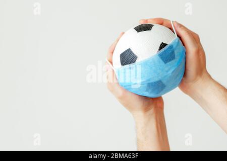 Soccer ball in medical protective mask in Man's hands on light background during coronavirus with copy space. Soccer on pause due to coronavirus. Stock Photo