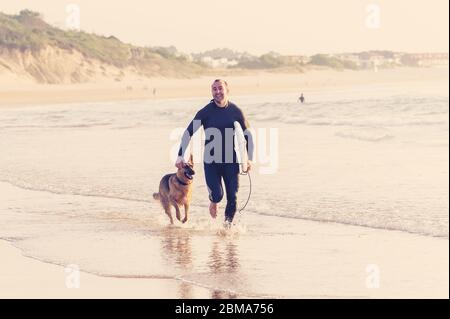 Surfer having fun with best friend german shepherd running and playing on dog-friendly beach at sunset. Summer fun surfing vacation with your dog, pet Stock Photo