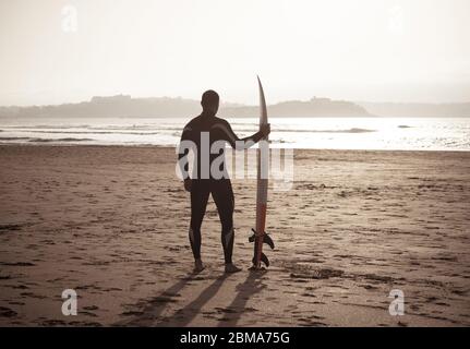 Rear view of strong surfer with surfboard on the beach at sunset or sunrise. Silhouette of surf man standing looking at ocean waiting for the high wav Stock Photo