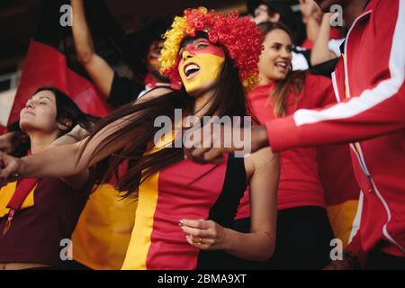 Woman in stadium wearing a wig and her face painted in german flag colors cheering for their national team. Female from Germany enjoying in fan zone. Stock Photo