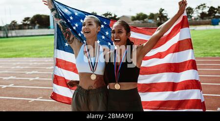 Female athletes on running track celebrating victory holding american flag. Sprinter carrying US flag and screaming after winning the sports event. Stock Photo