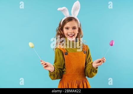 Photo of joyful blonde girl in toy rabbit ears smiling and holding colorful eggs isolated over blue background Stock Photo