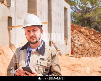 Caucasian bearded civil engineer or construction worker in white hardhat uses his pnone opposite construction site Stock Photo