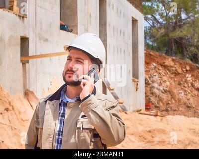 Caucasian bearded civil engineer or construction worker in white hardhat makes phone calls opposite construction site Stock Photo