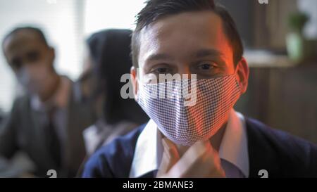 Positive businessman correcting protective mask on his face. Portrait of young man in office illuminated by neon light against background of people Stock Photo