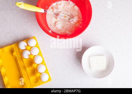 On the kitchen table is a red bowl with kneading dough from cottage cheese and with a whisk, a white bowl with butter and a dozen eggs in a yellow pla Stock Photo