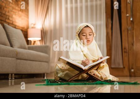 beautiful little girl reading quran wearing muslim hijab at home by herself Stock Photo