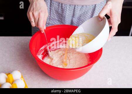 The girl holds a white cup in her hands with melted butter and pours it into whipped dough in red, stirring it with a spoon, eggs stand nearby. Cookin Stock Photo