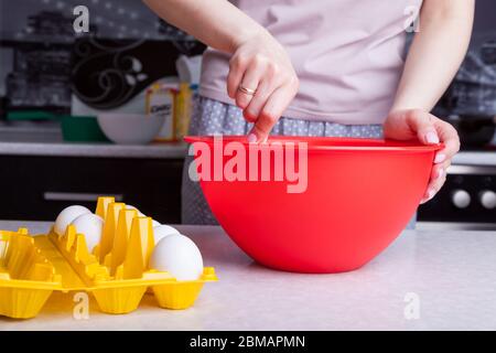 Female hands knead the ingredients in a red bowl in the kitchen for making dough and baking Easter cake at home, next to the table are eggs in a yello Stock Photo