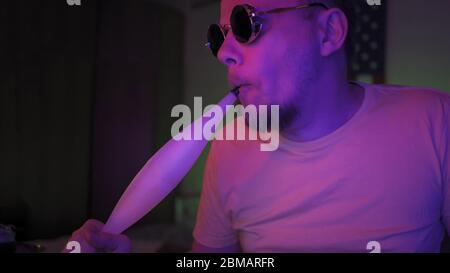 Man inhales the smoke of a hookah In Neon Lighting, Close Up Portrait of caucasian smoker in in dark room with fluorescent glowing. Quarantine at home Stock Photo