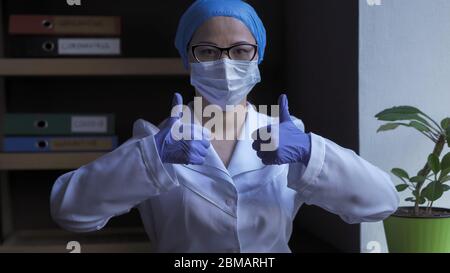 Doctor In White Uniform Shows Thumbs Up Sign. Female Medical Worker In Protective Mask And Disposable Gloves Shows Thumbs Up Gesture Giving Good Stock Photo