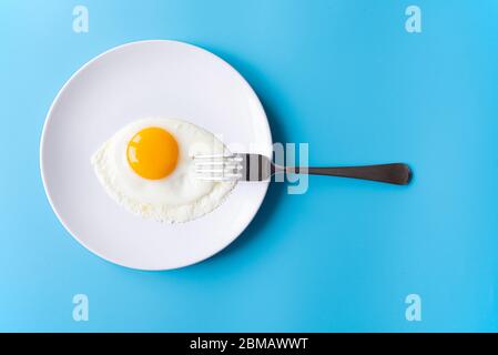 Breakfast, fried egg, yolk, omelet and fork on white plate with color table, creative picture Stock Photo