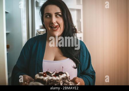 Plus size woman dreamily licks her lips while going to eat Dessert. Overweight woman took a chocolate cake with cherries from fridge Stock Photo