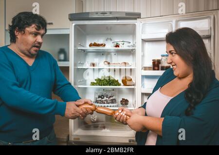 Plus positive couple fighting for sausages, hungry man and woman take sausages from each other standing near an open refrigerator with healthy food Stock Photo