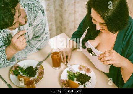 Body positive couple eats healthy food while sitting at kitchen table, man and woman eat salad and fruits cut on their plates Stock Photo