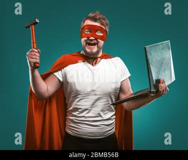 Smiling Super hero man holds hammer and laptop ready to repair the computer. Man In Red Super Hero Uniform posing on biscay green background. Computer Stock Photo