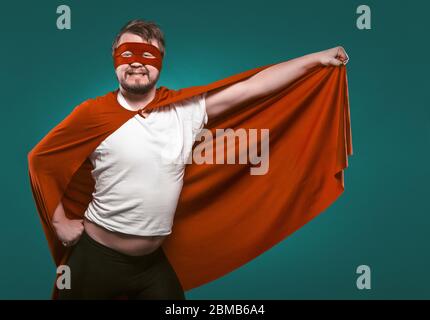 Funny Super Hero Man Ready To Fly Save World. Smiling Man In Mask And Red Superhero Costume Holding His Cloak In His Hand Posing On Biscay Green Stock Photo