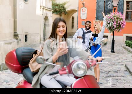 Pretty cheerful brunette Caucasian girl sitting on the red scooter and using phone, while her multiracial friends having fun and making selfie photo Stock Photo