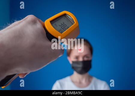 Man hand holding yellow pyrometer and measuring temperature of woman in medical face mask looking at camera: close up view, selective focus Stock Photo