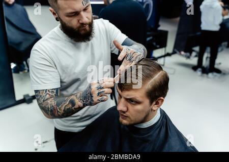 Attractive male is getting a modern haircut in barber shop. Tattooed Barber wets hair by spray and combs them. Stock Photo