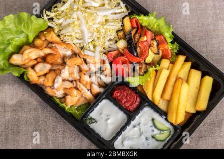 Traditional doner kebab in a black plastic container, sour cream, fried potato, roast meat, sauces, salad, cabbage and roasted vegetables on grey back Stock Photo