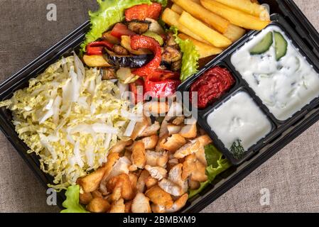 Traditional doner kebab in a black plastic container, sour cream, fried potato, roast meat, sauces, salad, cabbage and roasted vegetables on grey back Stock Photo