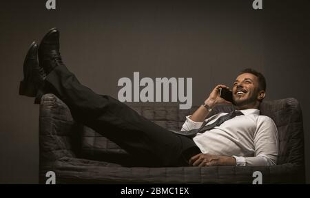 Businessman with mobile lies throwing legs on the sofa. Smiling man lying at couch cross-legged talking over smartphone on grey back. Business Stock Photo