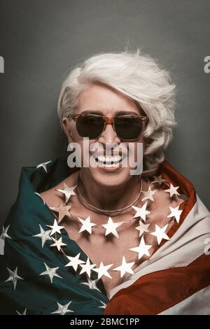 American patriotic woman celebrate Independence Day of USA. Senior woman laughs looking at camera wrapped in american flag wearing beads of stars Stock Photo