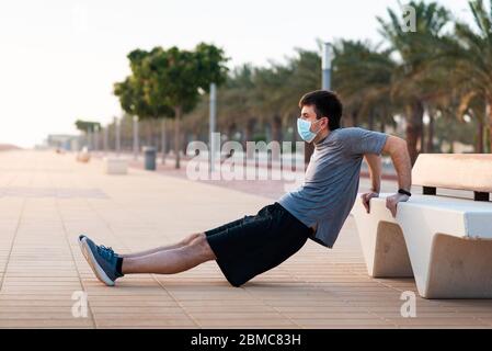 Man doing triceps dips on a bench outdoors and wearing protective surgical face mask Stock Photo