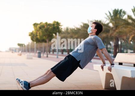 Man doing triceps dips on a bench outdoors and wearing protective surgical face mask