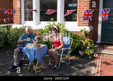 As the Coronavirus lockdown continues over the May Bank Holiday, the nation commemorates the 75th anniversary of VE Day (Victory in Europe Day, the day that Germany officially surrendered in 1945) and in Dulwich, neighbours and residents emerge from their homes to party while still observing social distancing rules. A couple enjoy afternoon tea and cake beneath Union Jack bunting outside their home, on 8th May 2020, in London, England. Stock Photo