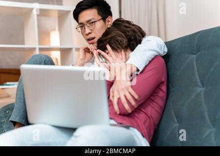 Photo of scared multinational couple hugging and using laptop while sitting on sofa at home Stock Photo