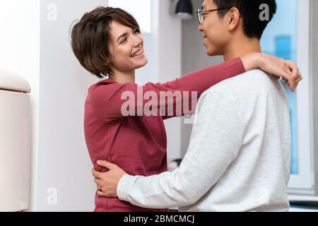 Photo of romantic multinational couple laughing and hugging while dancing in kitchen at home Stock Photo