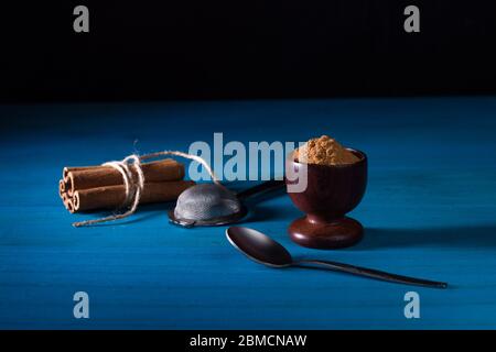 Ground cinnamon powder in wooden cup, a pack of cinnamon sticks, a little spoon and metal sifter on blue wooden table with dark background. Ceylon cin Stock Photo