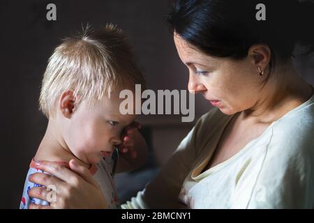 Tired mother hugging and calming little cute sad upset crying caucasian blond toddler boy at home indoor. Mom holding sleepy sick son. Child health Stock Photo