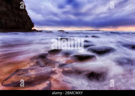 ROcks on sandy beach of Sydney Northern beaches near tall headland cliff at sunrise during stormy weather.