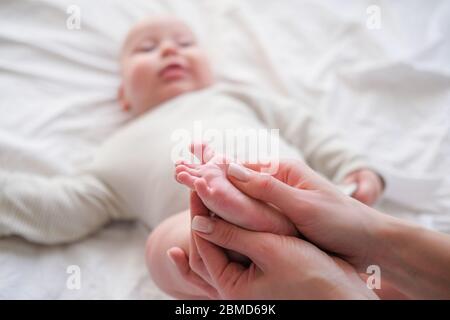 Baby feet in mother hands. Young caucasian woman makes massage for happy infant baby on white bed at home. Babycare, sport and happy motherhood. Stock Photo