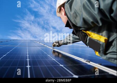 Electrician in safety helmet, uniform and working gloves, setting a shiny new solar battery with help of hex key, blue sky on background. Concept of alternative green energy sources and innovations. Stock Photo
