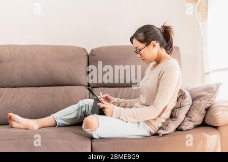 Young Asian woman working from home due to coronavirus pandemic lockdown. Graphic designer using tablet for work at home lying on couch Stock Photo