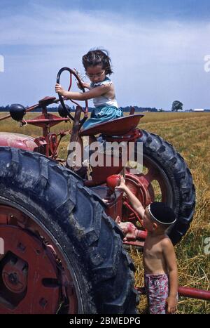 Two children playing in the sunshine on a tractor on the family farm, USA in the early 1950s. Stock Photo