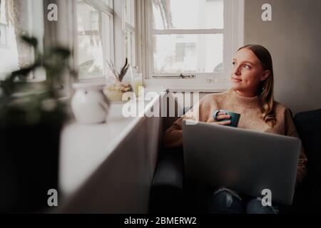 Thoughtful young businesswoman working on laptop computer sitting at home holding a coffee cup in hand looking outside window Stock Photo