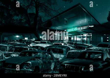 Post apocalyptic landscape. Abandoned, rusting cars gathered in front of a drive in still with its green neon sign still glowing. Stock Photo