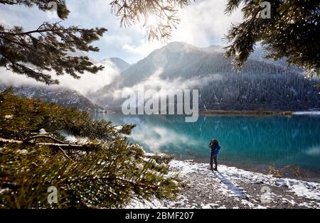 Tourist with backpack and camera taking picture at the snow beach near mountain lake in Kazakhstan, Central Asia Stock Photo