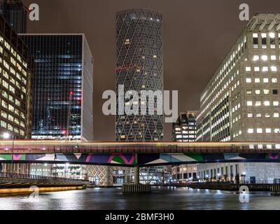 London, England, UK - March 10, 2020: A DLR train leaves light trails as it crosses Middle Dock among the office blocks and skyscrapers of London's re Stock Photo