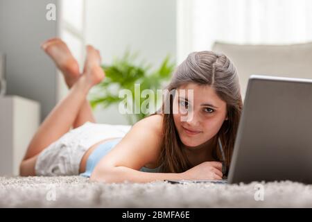 Young woman lying on a bed with her laptop computer relaxing and looking thoughtfully at the camera in a low angle view Stock Photo