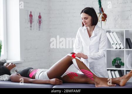 Physiotherapist applying kinesiology tape to patient knee.Therapist treating young female African American athlete. Kinesiology taping. Post traumatic Stock Photo