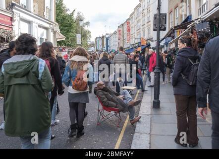 Crowds of shoppers at Portobello Market with sleepy man in chair watching his shop. Kensington & Chelsea, Portobello Road, Notting Hill, London, UK Stock Photo