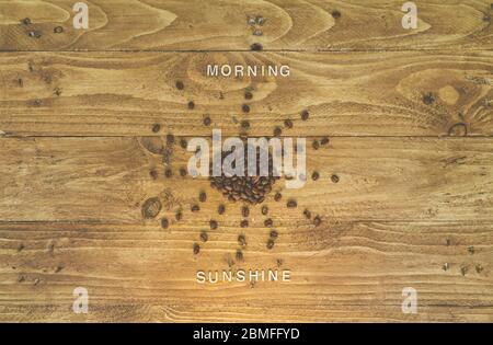 Coffee beans shaped as the sun on a wooden background, with the words “morning sunshine” written out with wooden letters Stock Photo