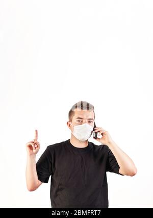Young man in face mouth nose virus mask pointing finger above him as he talks over mobile phone, isolated on white - empty space for text. Can be used Stock Photo