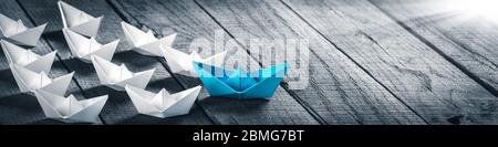 Blue Paper Boat Leading A Fleet Of Small White Boats On Wooden Table With Sunlight - Leadership Concept Stock Photo